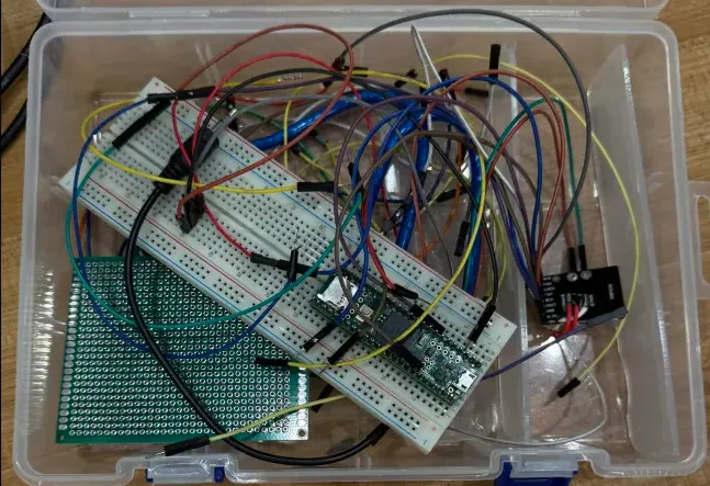a plastic case holding a breadboard wired with jumper wires, a microcontroller, and a small audio board